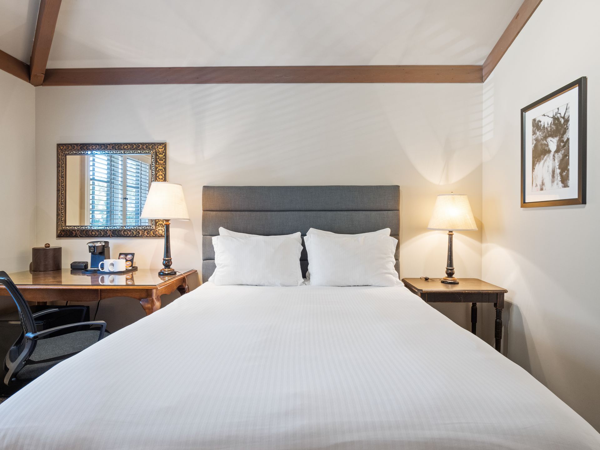 A hotel room featuring a bed with white bedding, a gray headboard, two lamps, a desk with a chair, and wall art.