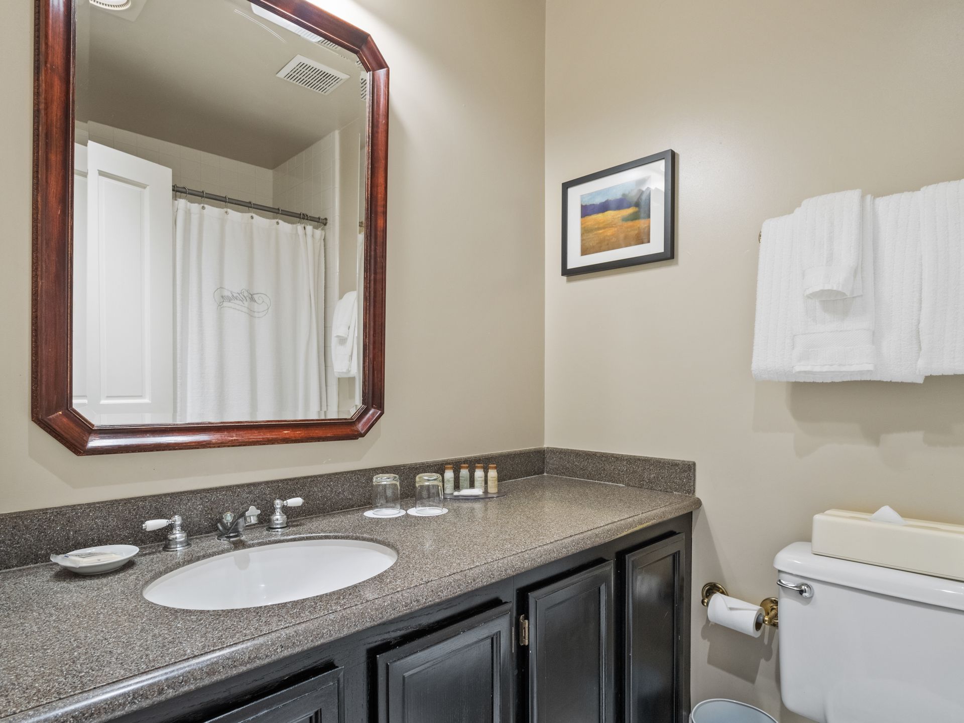 A hotel bathroom with a granite vanity, dark wood cabinets, a large mirror, and white towels hanging on the wall.