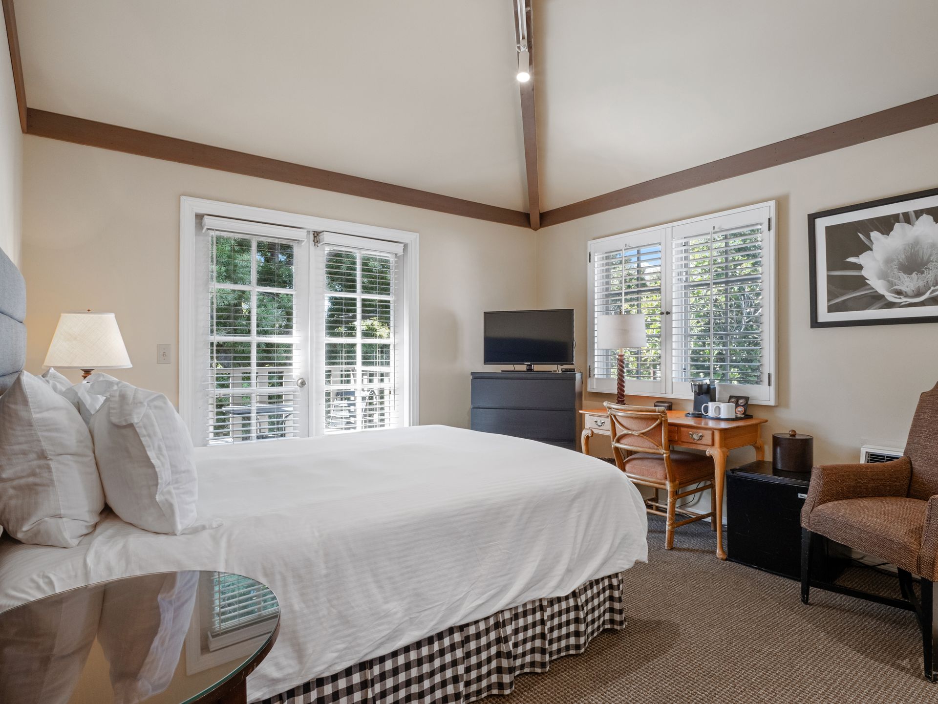 A hotel room featuring a queen bed, a desk with a television, a small armchair, and glass doors leading to a balcony.