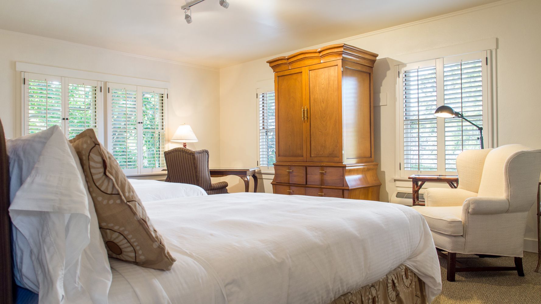 A well-lit hotel bedroom features a large bed, a wooden wardrobe, two white wingback chairs, and plantation shutters.