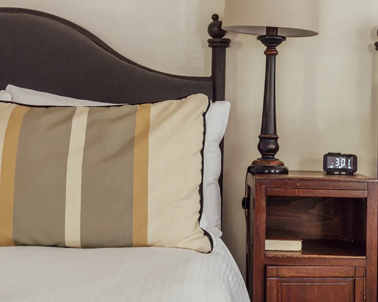 A striped pillow on a white bed next to a wooden nightstand with a lamp and a digital alarm clock.