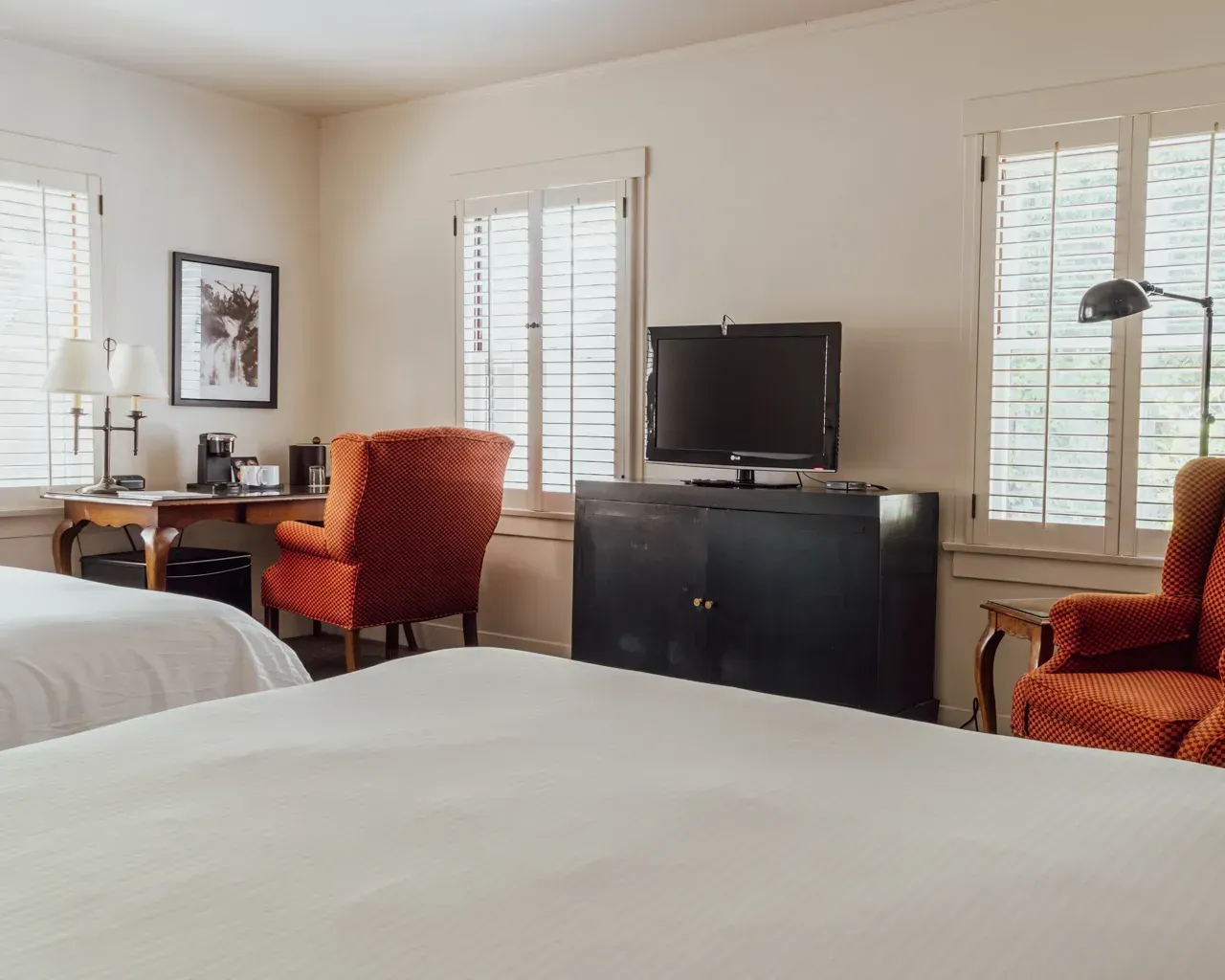 A hotel room with two white beds, two orange-patterned armchairs, a wooden desk, and a television on a black cabinet.