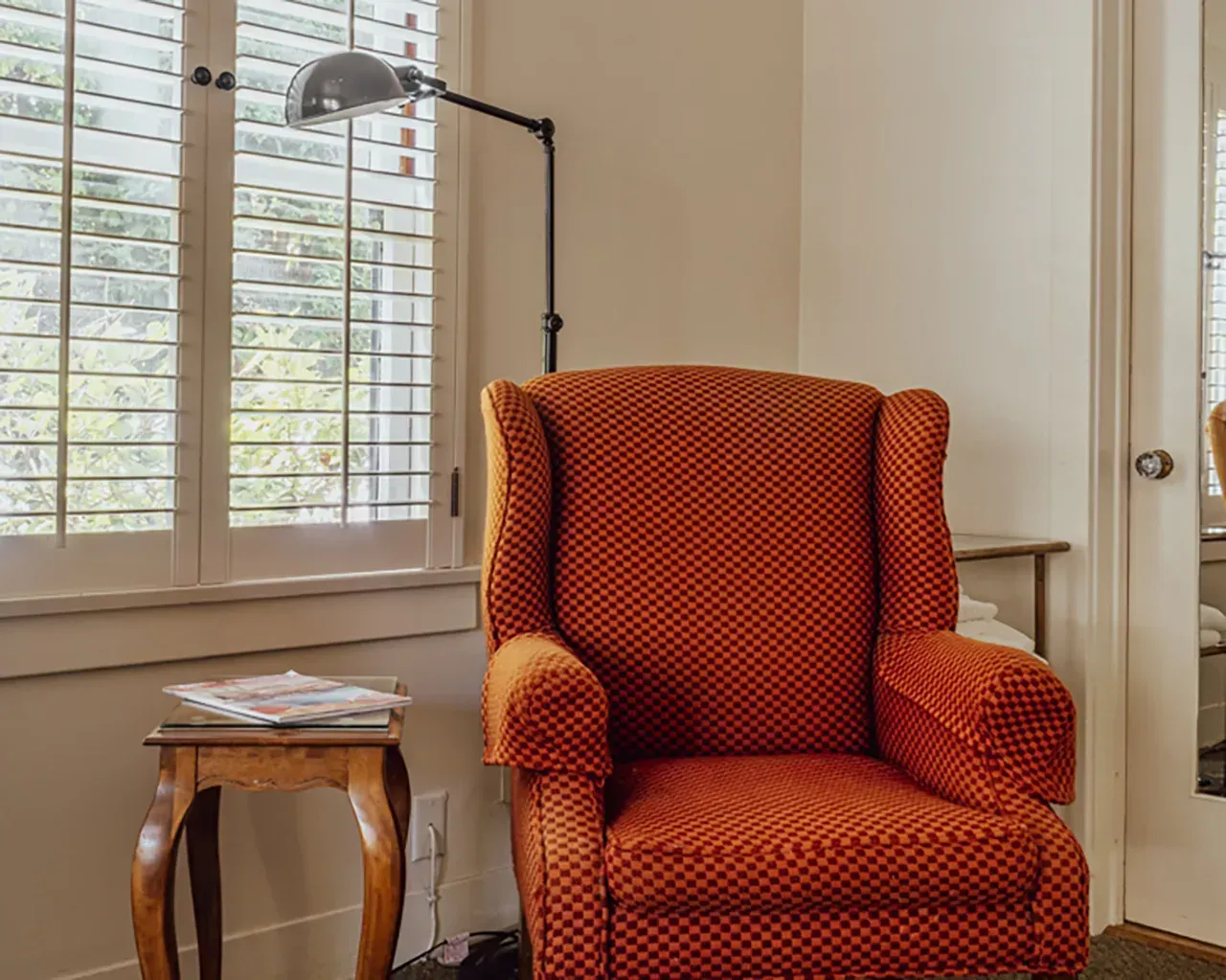 An orange patterned wingback chair sits beside a small wooden side table with a magazine under a black reading lamp.