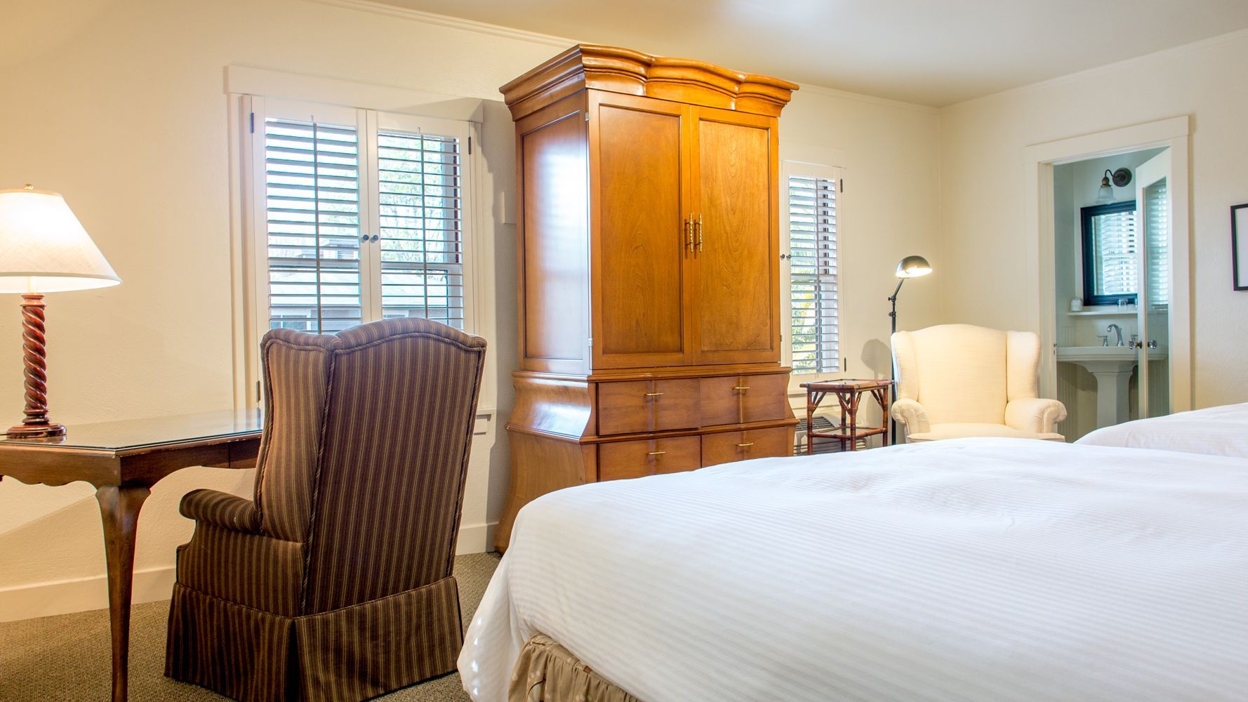 A bright bedroom with a white-clad bed, a large wooden armoire, a striped armchair at a desk, and a cream-colored chair.