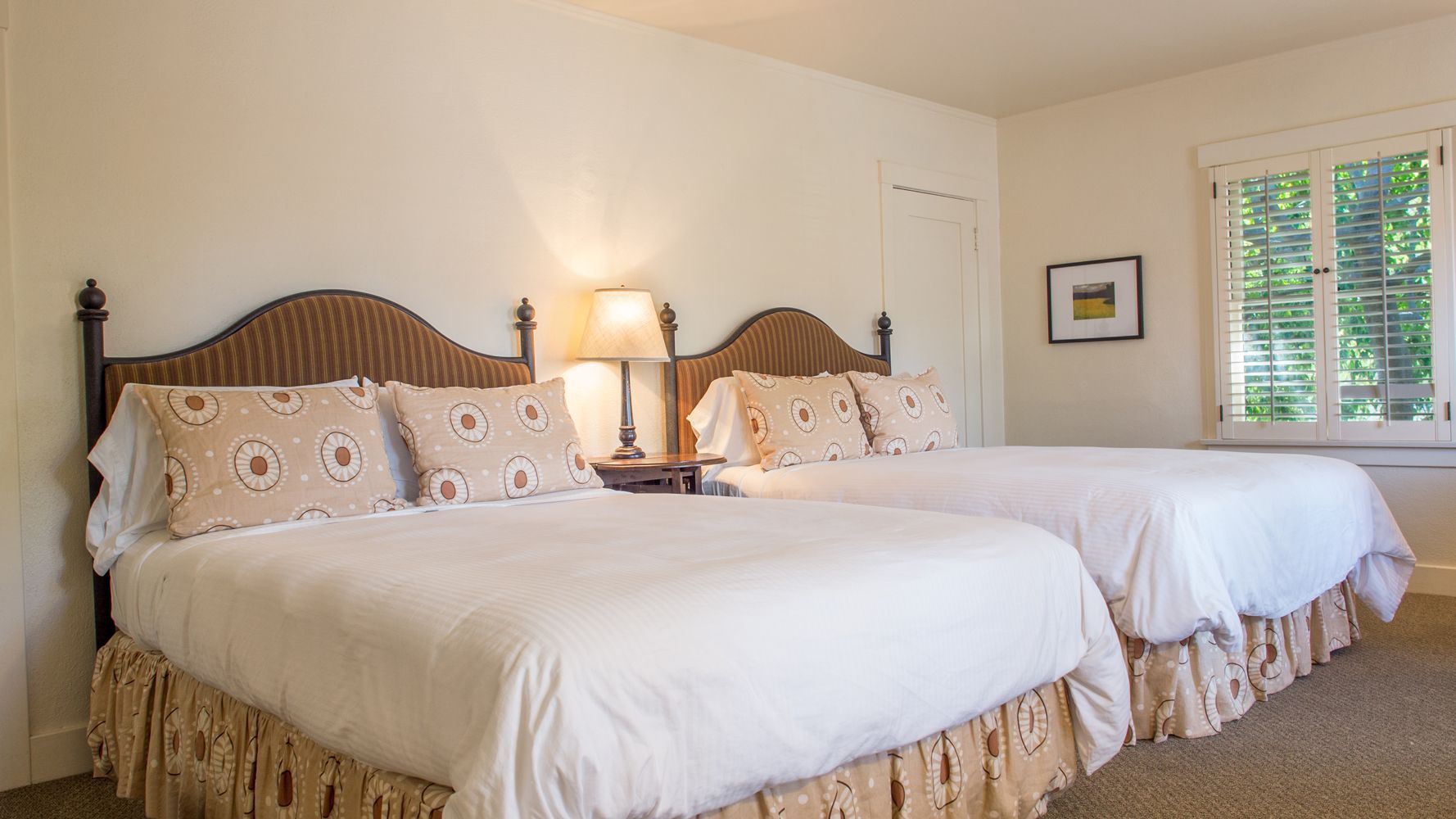 A hotel room with two queen beds, wicker headboards, beige bedding, a lamp on a side table, and a window with shutters.