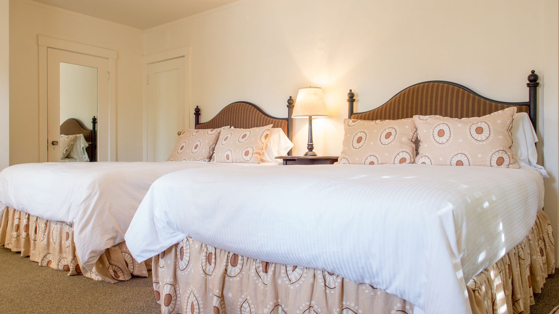 Two beds with white linens and patterned beige headboards and bed skirts, separated by a nightstand with a lamp.