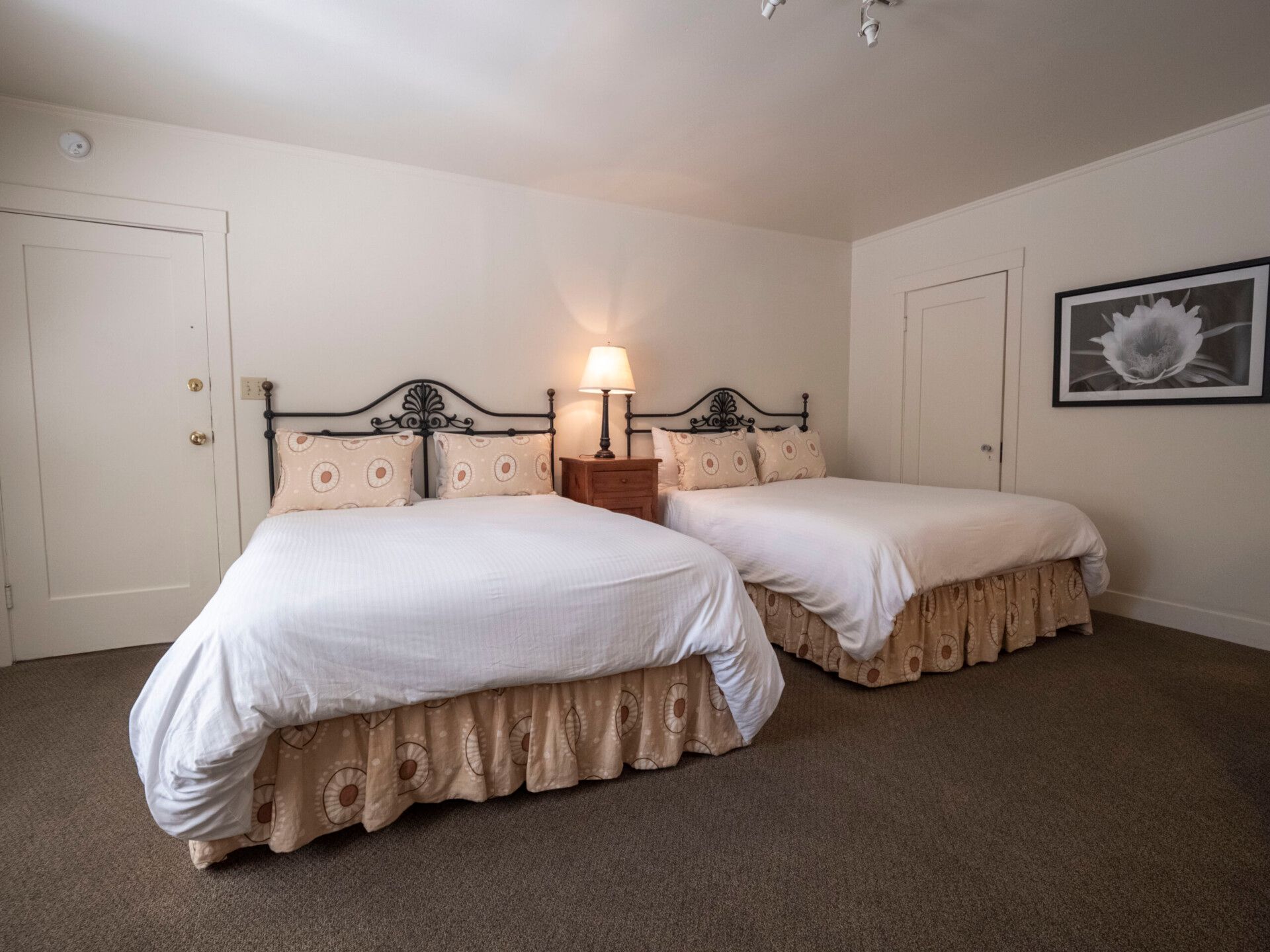 A hotel room with two queen beds, each with white linens and patterned beige bed skirts, separated by a nightstand.