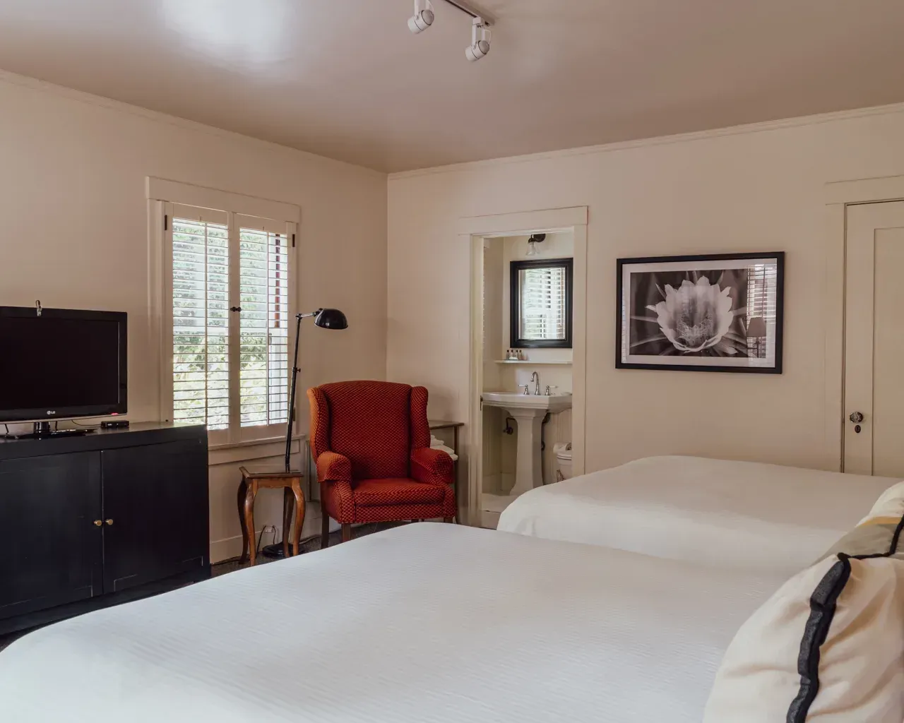 A hotel room with two white beds, a red armchair, a black media console with a TV, and an open bathroom with a pedestal sink.