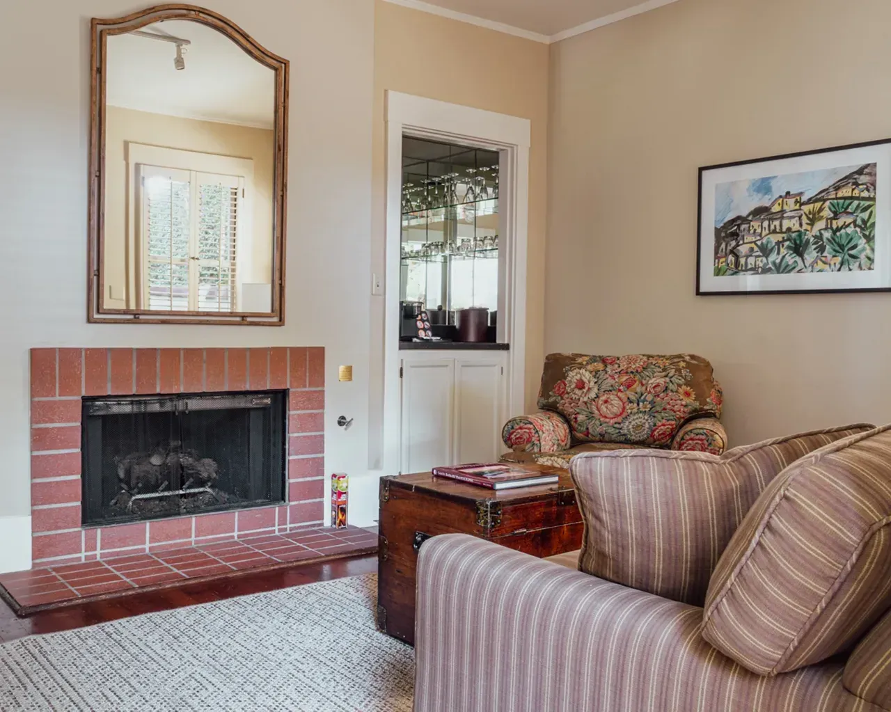 A cozy room featuring a brick fireplace, a mirror, a patterned armchair, and a striped sofa on a light textured rug.