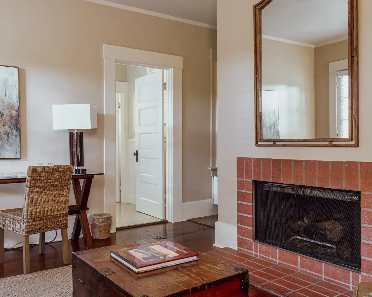 A cozy room featuring a brick fireplace, a wooden chest coffee table, a wicker desk chair, and a large wall mirror.