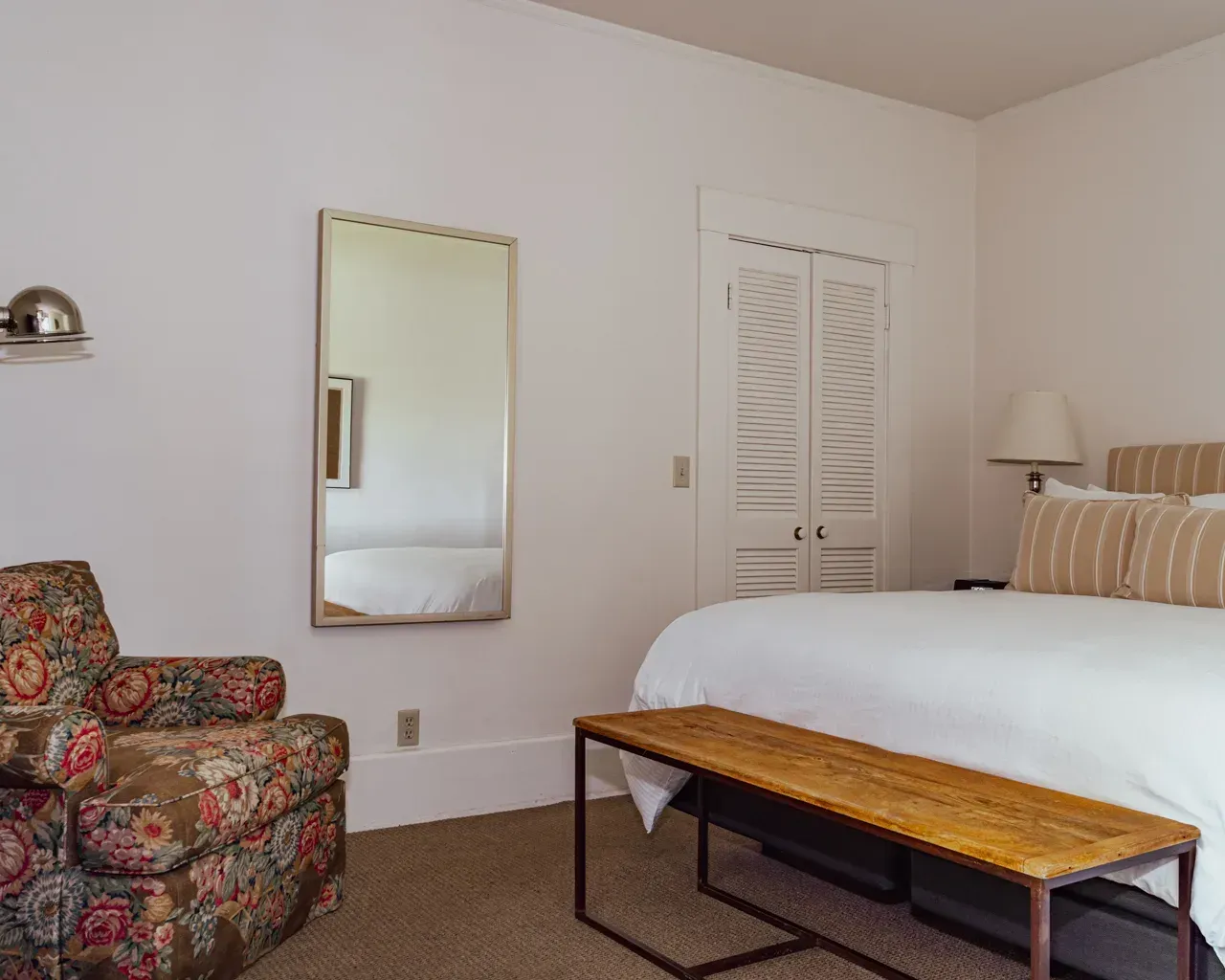 A bedroom features a floral armchair, a wall mirror reflecting the bed, a wooden bench, and a bed with striped pillows.
