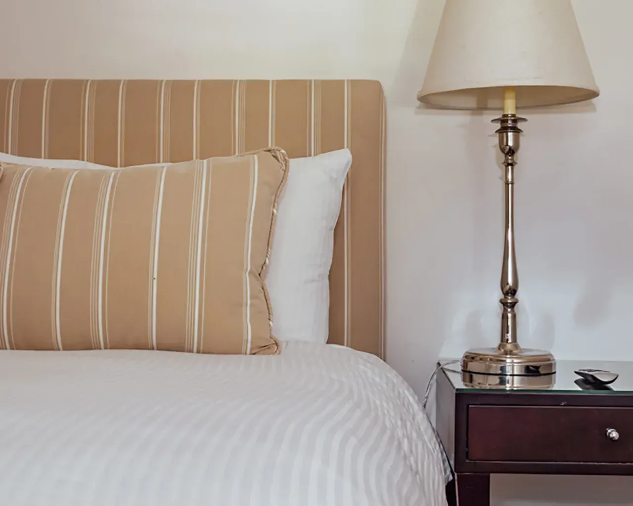 A tan striped headboard and matching pillow sit on a bed beside a bedside lamp on a dark wood table.