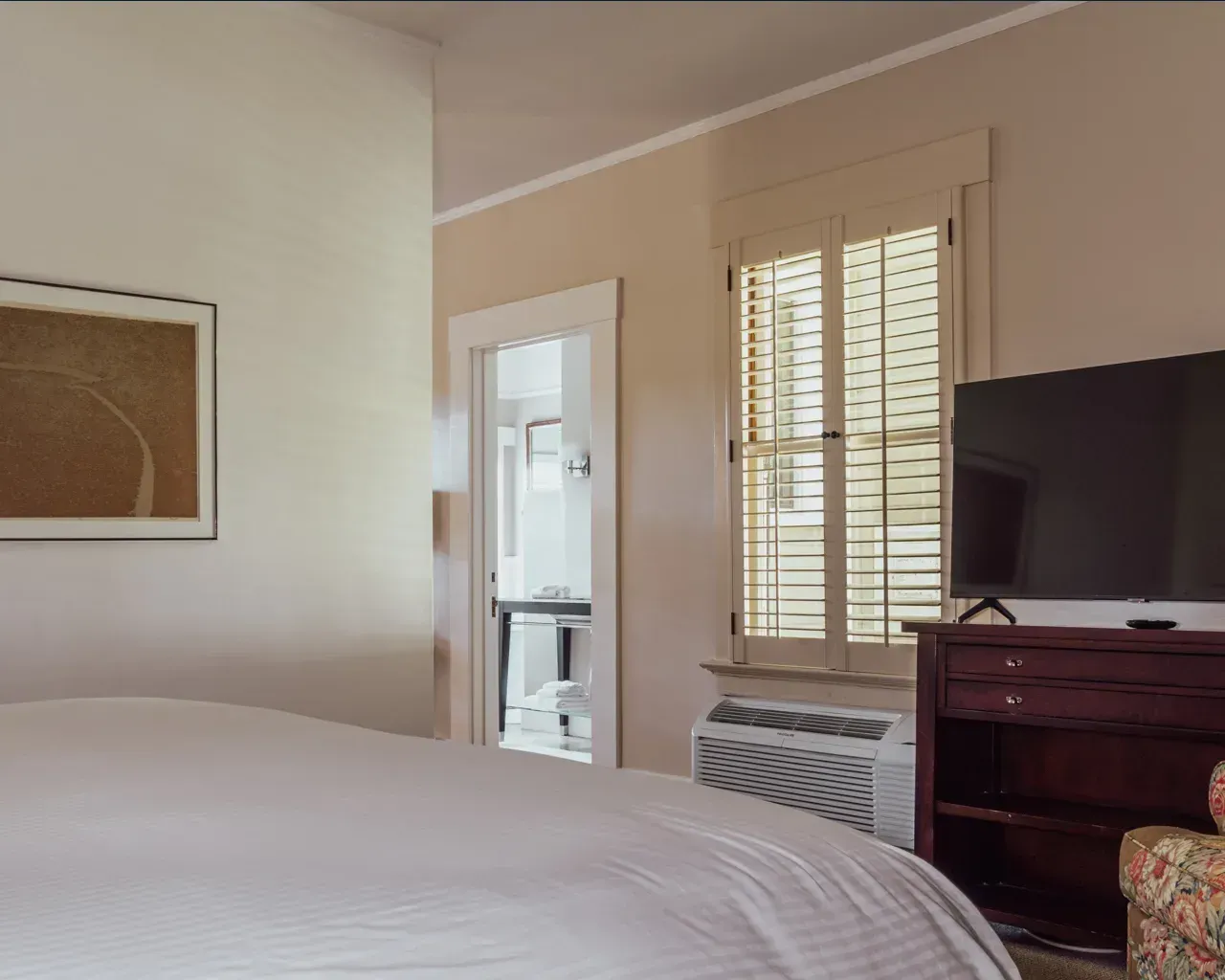 A bedroom interior with a white bed, a wall picture, a doorway to a bathroom, shuttered windows, and a TV on a dresser.
