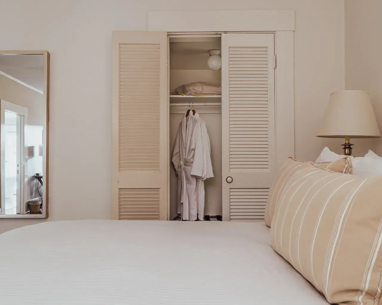 A beige bedroom interior featuring a white-made bed, a white robe hanging in an open louvered closet, and a table lamp.