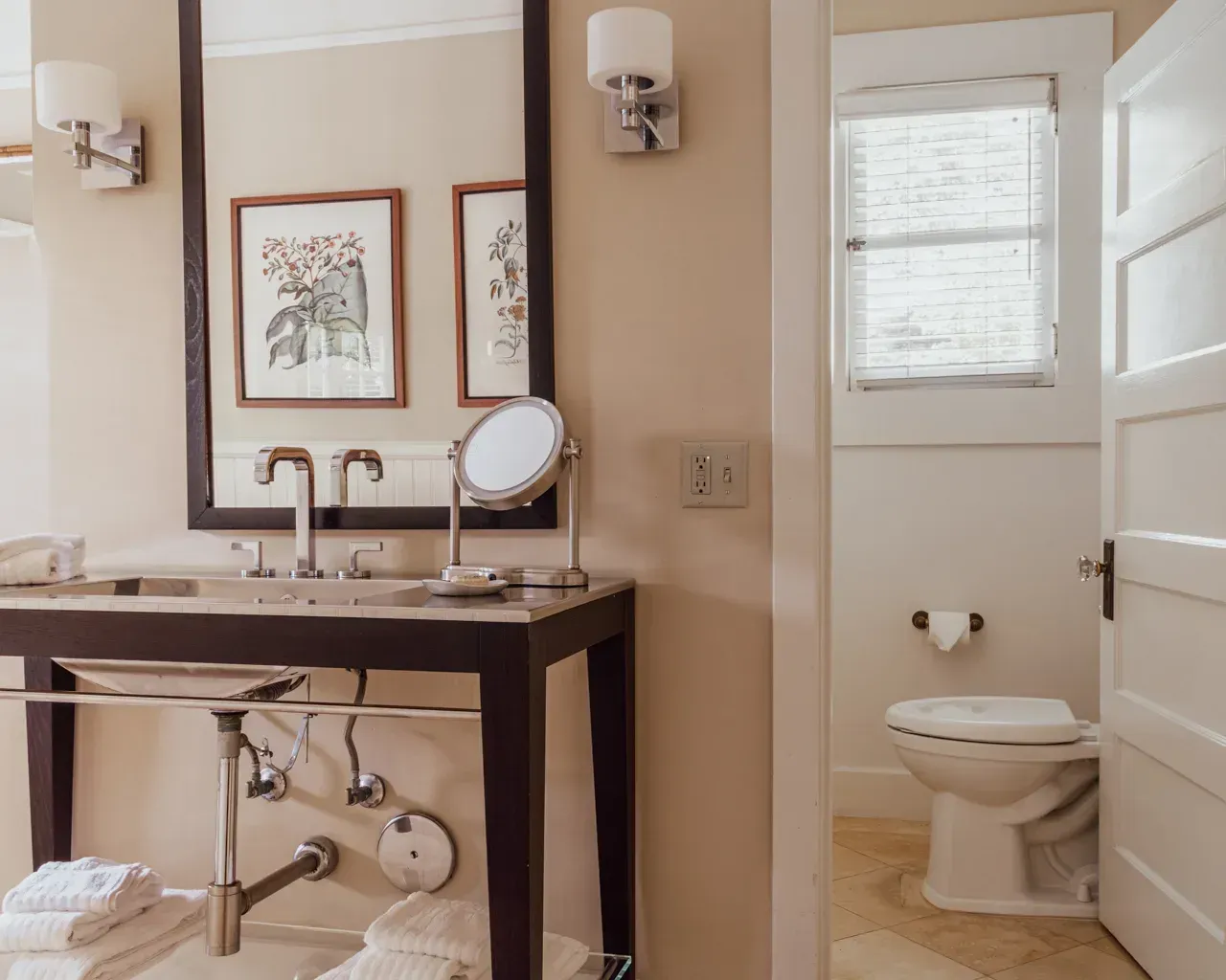 A bathroom with a dark-framed console vanity, a round mirror, and a small vanity mirror, opening into a separate toilet.