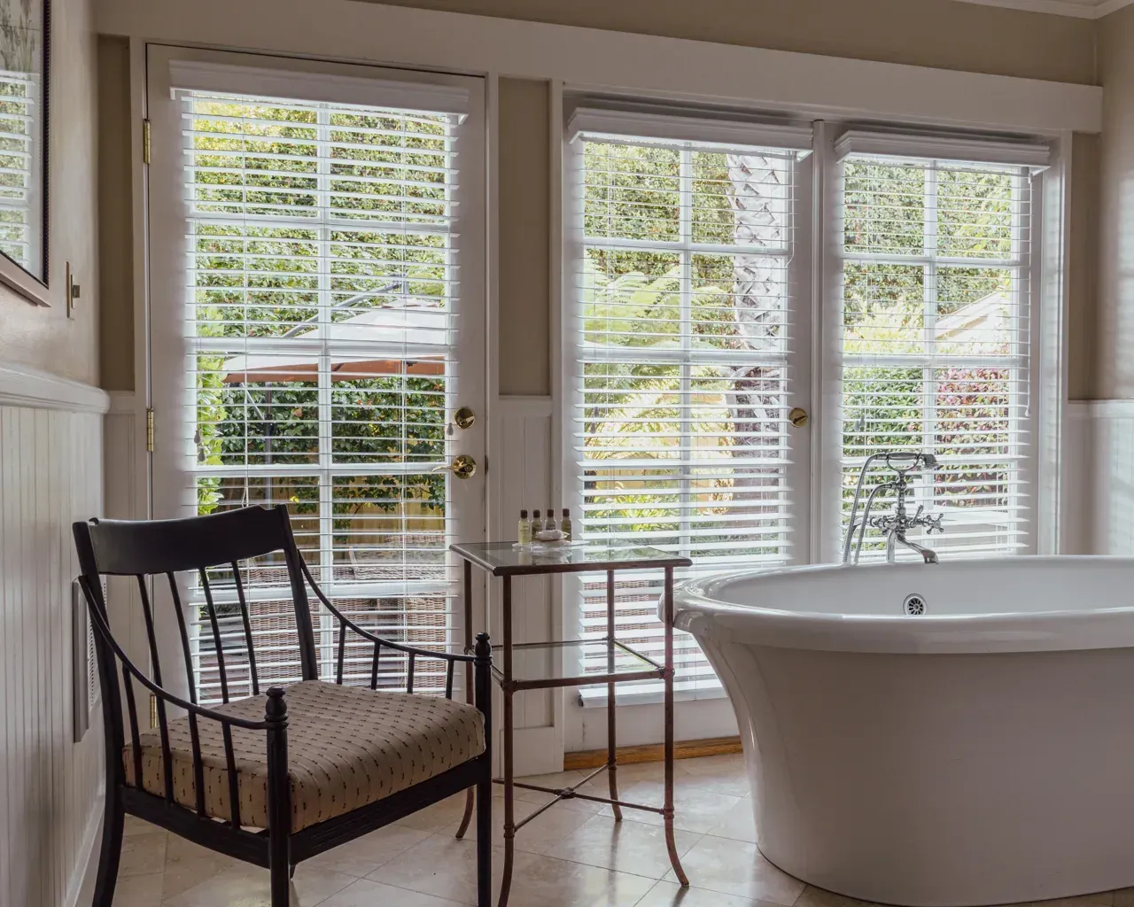 A freestanding white bathtub sits next to a small metal table and a dark wooden chair in a sunlit bathroom with glass doors.