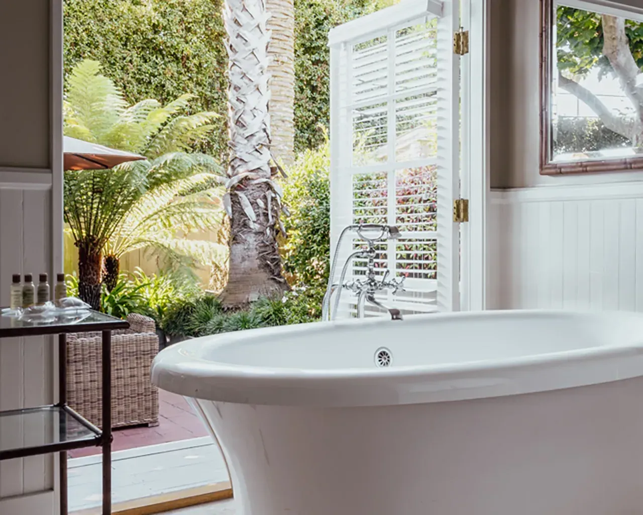 A white clawfoot bathtub in a bathroom, situated in front of a wide-open door looking out onto a lush, tropical garden.