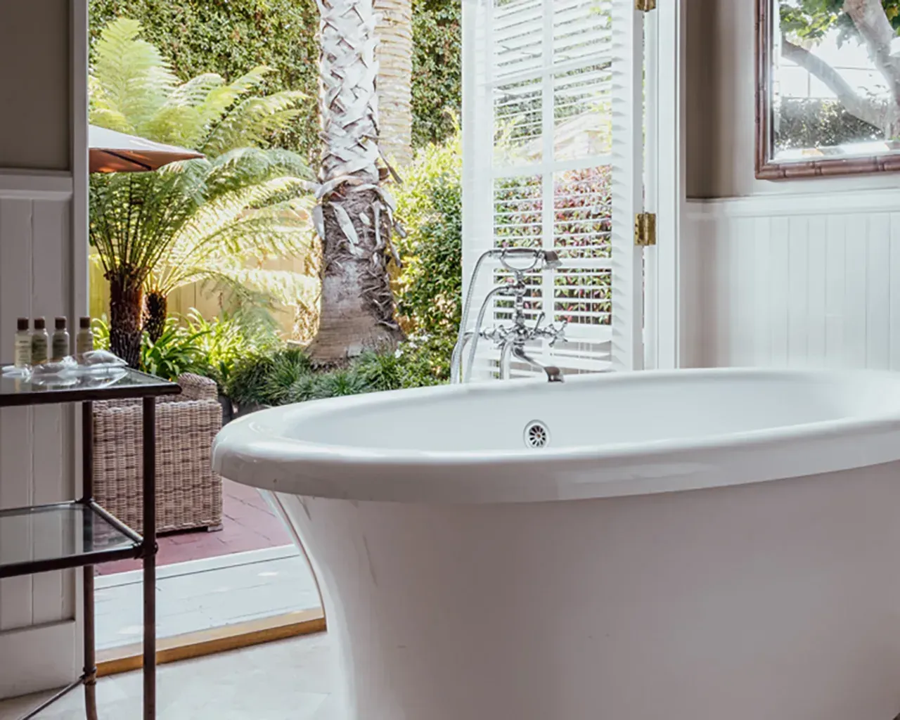 White freestanding bathtub in a bright, modern bathroom looking out toward a lush garden through open French doors.