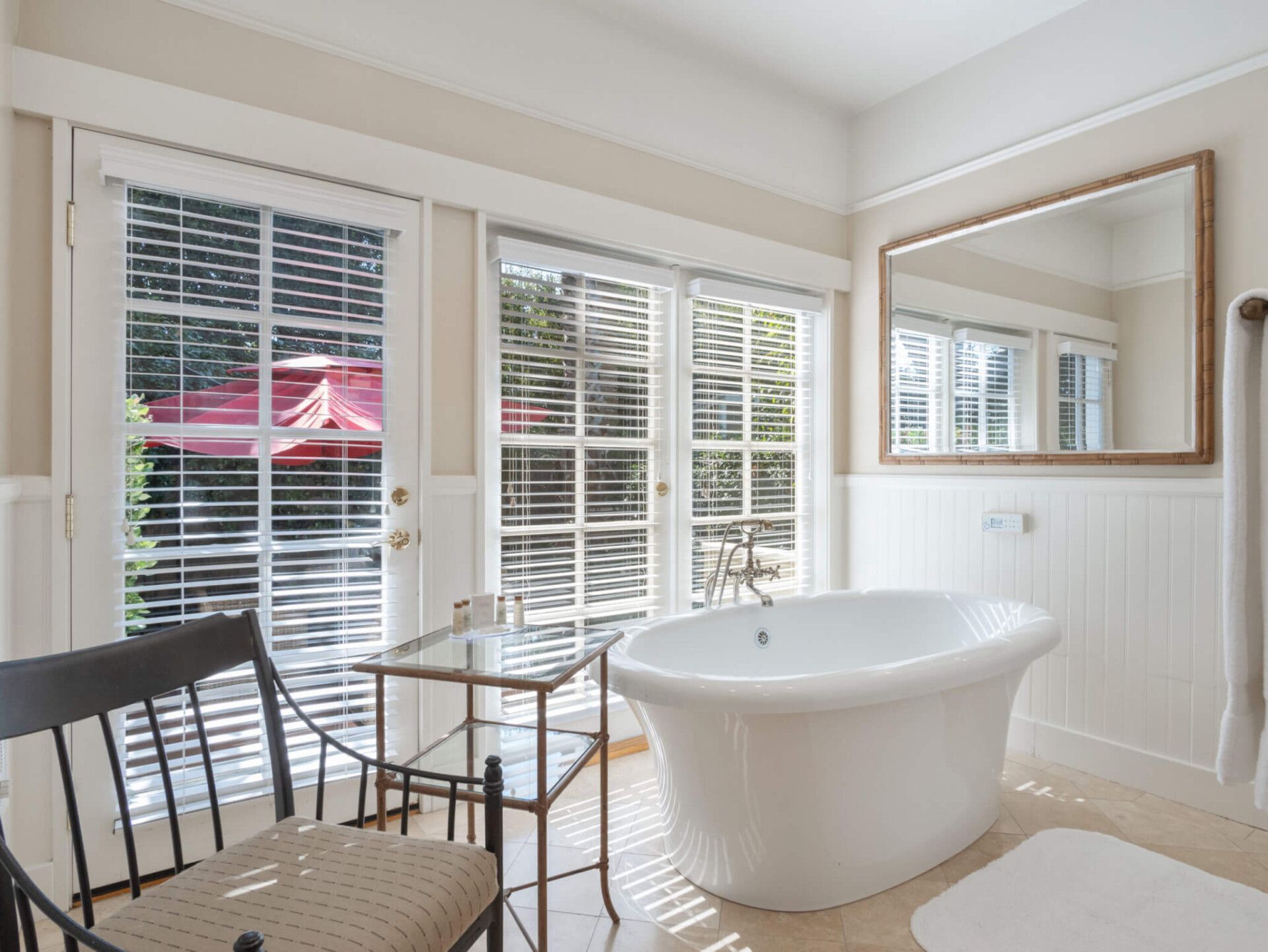A white freestanding bathtub in a bright, modern bathroom with glass doors, a side table, and a large wall mirror.