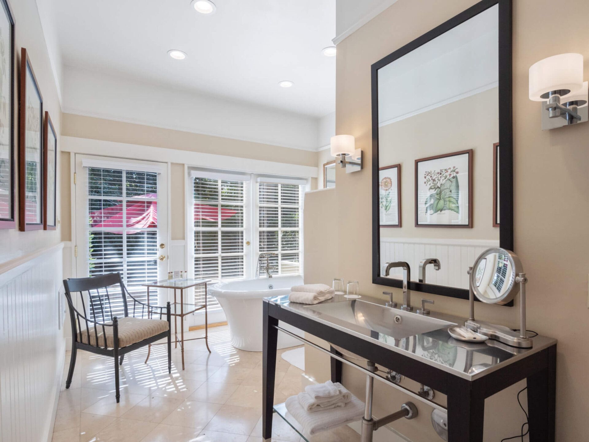 A bright bathroom with a black vanity, modern sink, large mirror, white soaking tub, and a chair near a windowed door.