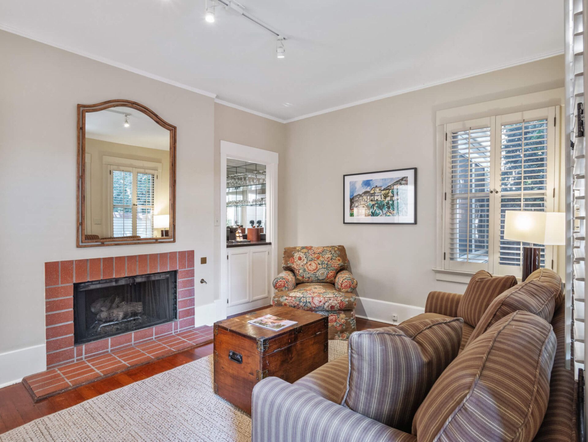 A living room featuring a red brick fireplace, a wooden chest coffee table, striped sofa, floral armchair, and a mirror.