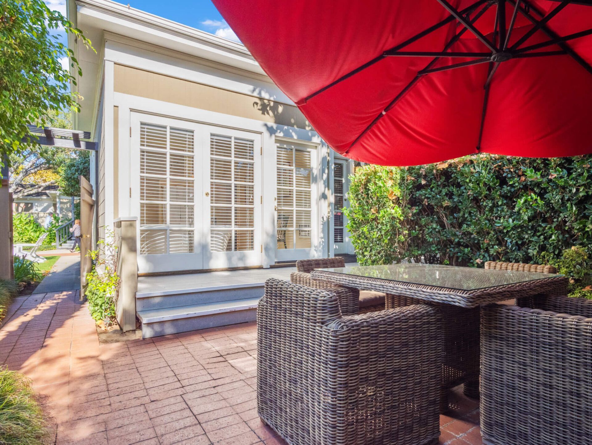 A patio with a wicker dining set under a large red umbrella, next to a building with glass French doors.
