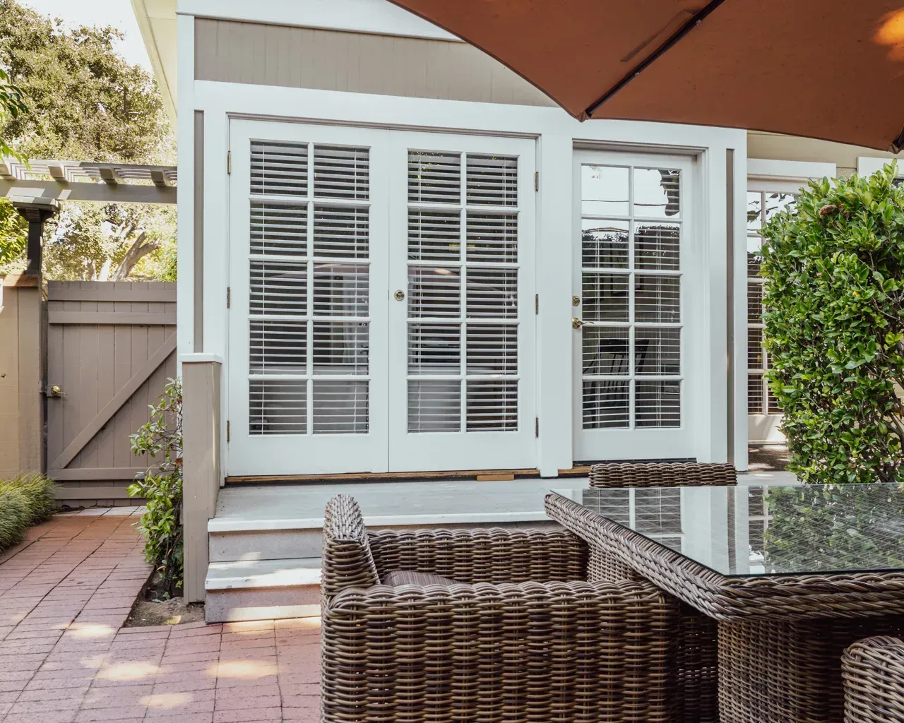A sunny patio with a wicker dining set and a large umbrella in front of a white house with French doors.