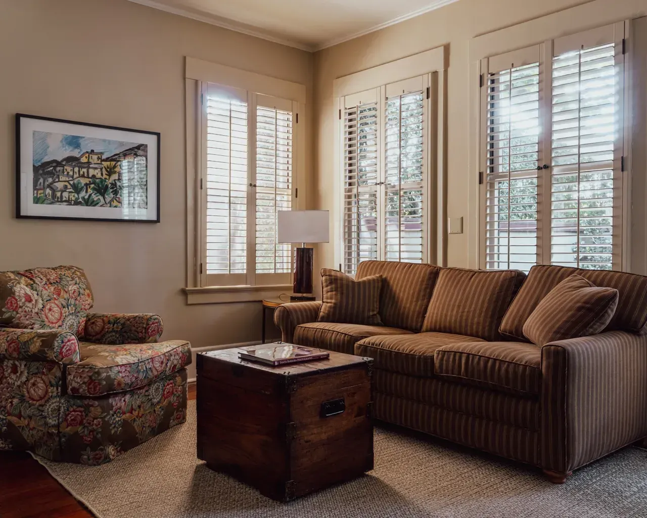 A cozy living room with a brown striped sofa, floral armchair, wooden trunk coffee table, and plantation shutters.