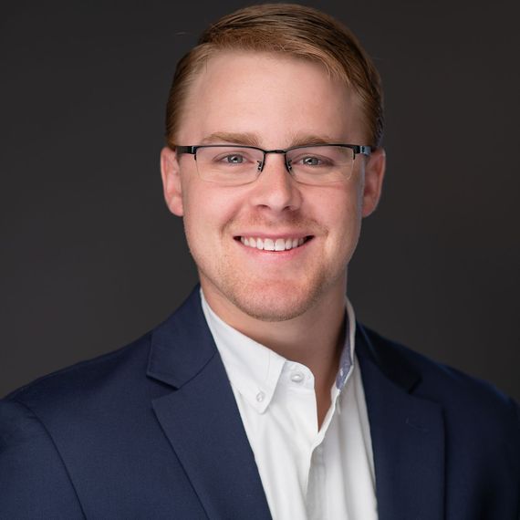 Smiling person in glasses wearing a navy blazer and white shirt against a dark background
