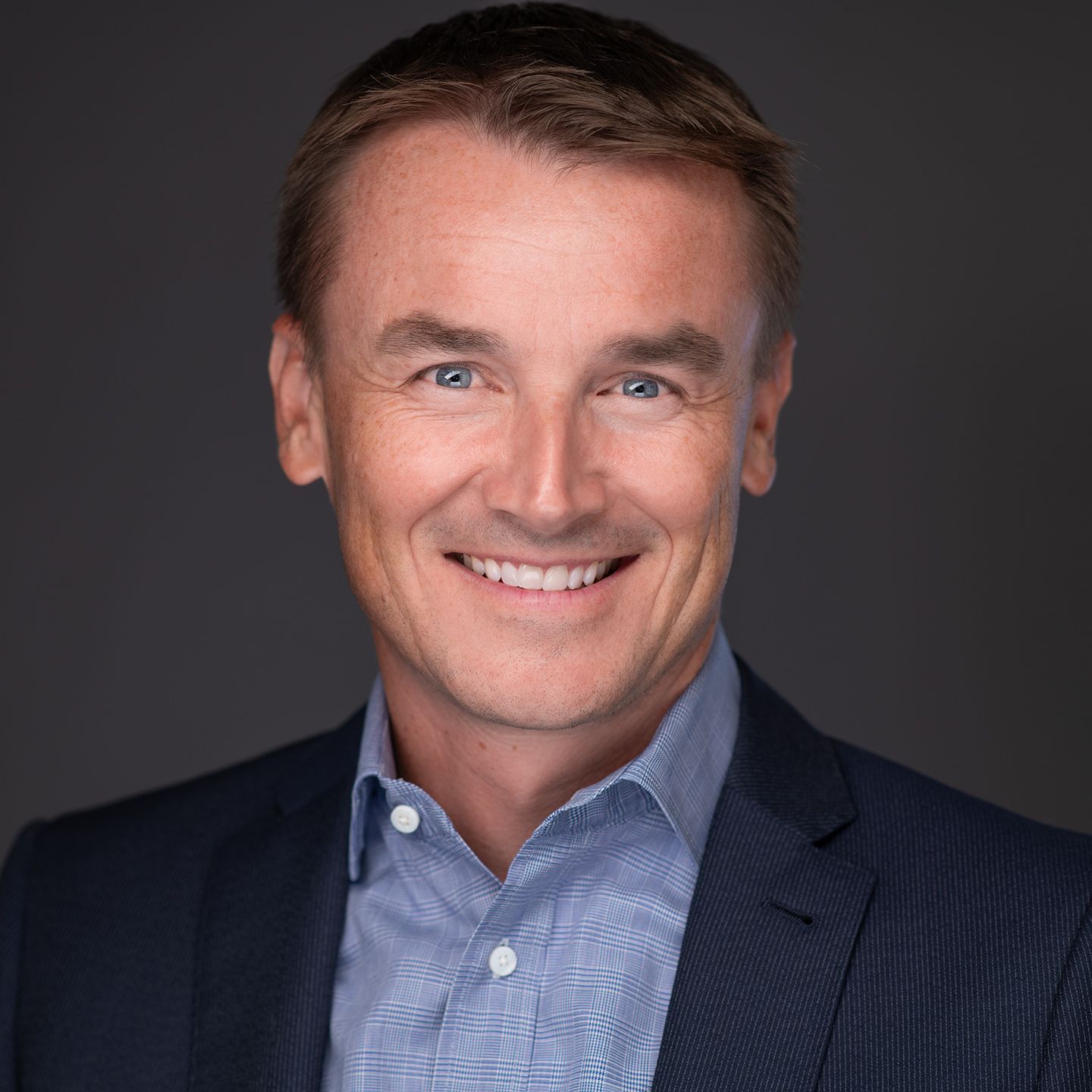 Smiling man in a navy blazer and blue shirt against a dark gray background