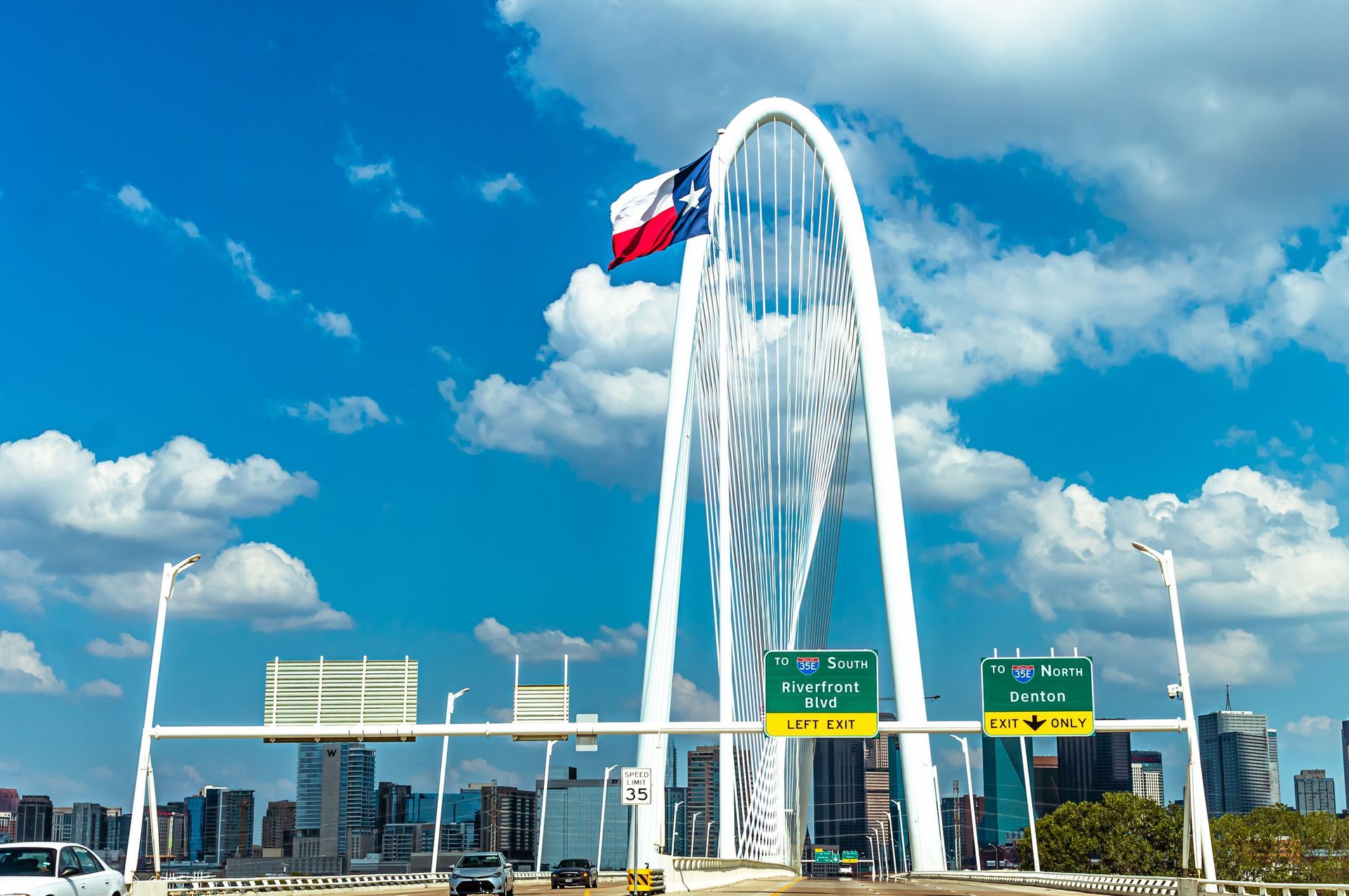 Suspension bridge with a white arch tower and a Texas flag against a blue sky