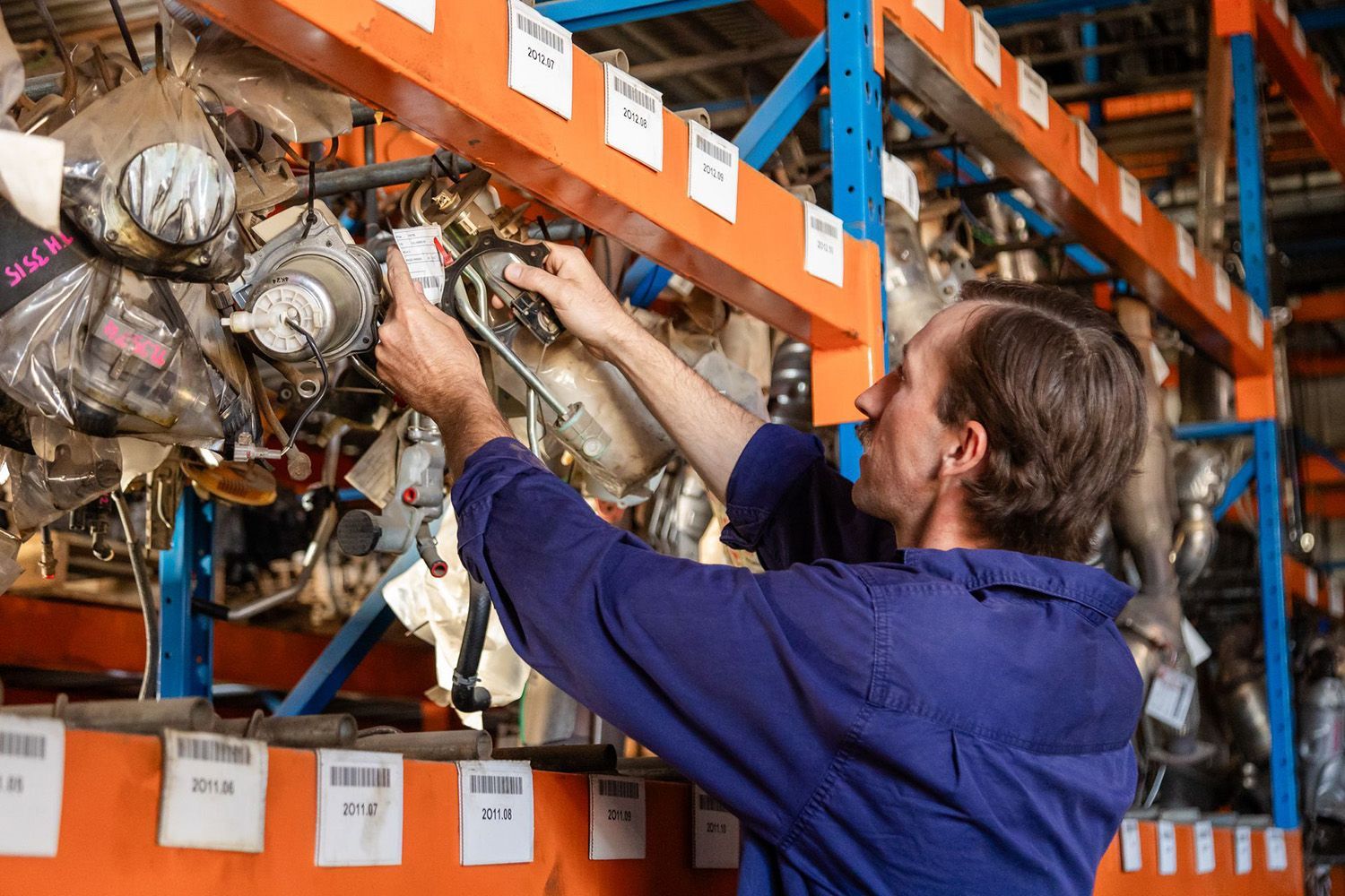 A Worker In A Blue Uniform Selects An Automotive Part From Shelves In A Storage Warehouse