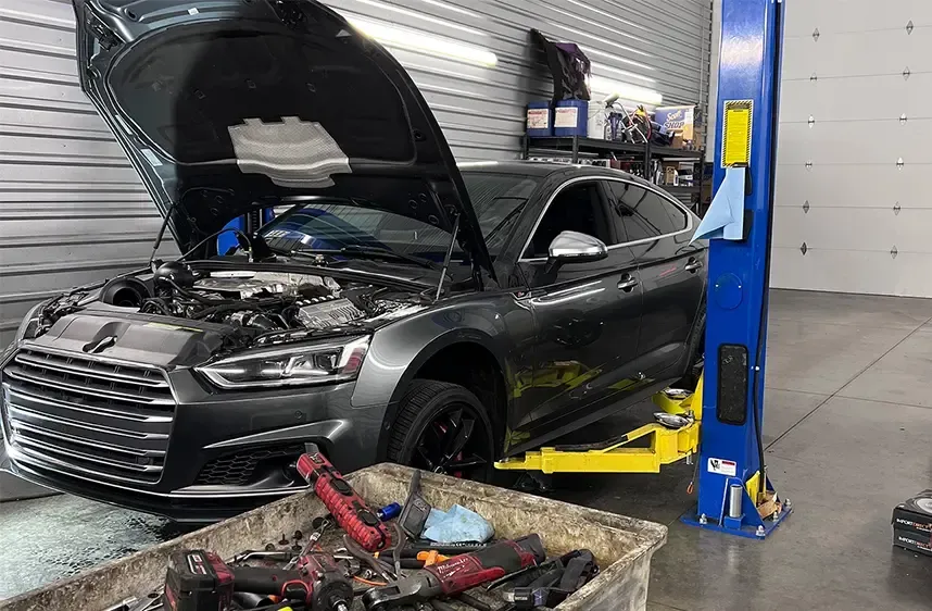 Gray car with open hood on a lift inside a garage; tools in a bin in front. | Mike's Garage CC