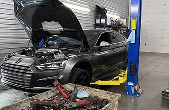 Gray car with open hood on a lift inside a garage; tools in a bin in front. | Mike's Garage CC
