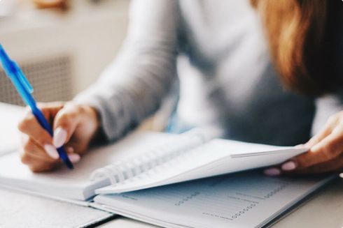 Hands writing in a notebook with a blue pen at a desk with papers nearby
