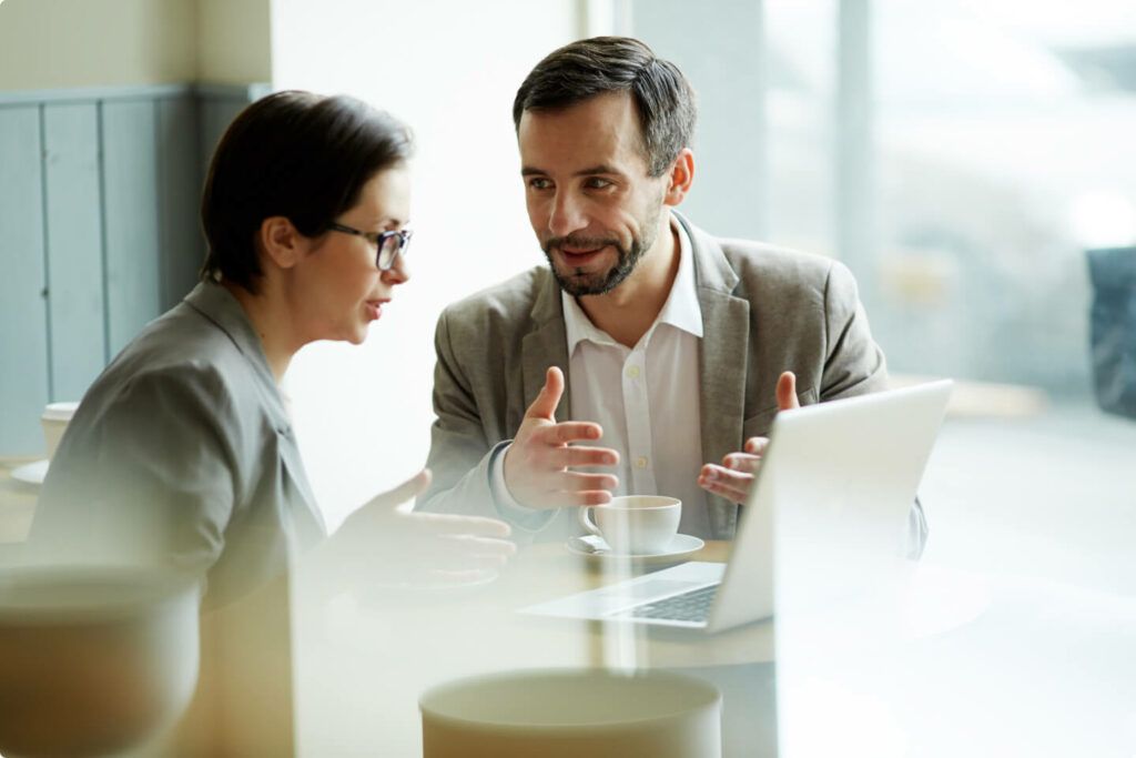 Two coworkers in a bright office discussing at a table with a laptop and papers