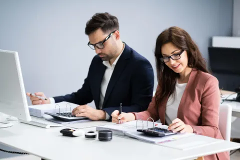 Two colleagues reviewing documents at a desk beside a computer in a bright office