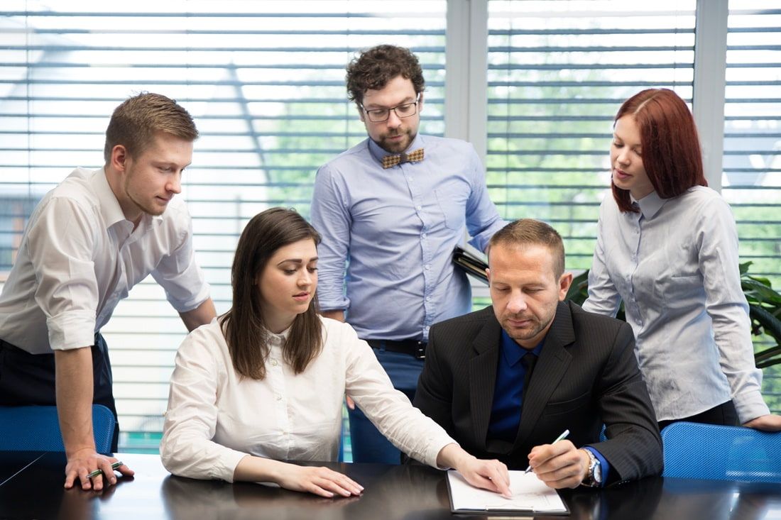 Five coworkers review a document together at a desk in a bright office.