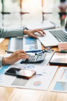 Hands using a calculator beside a laptop and financial charts on a desk