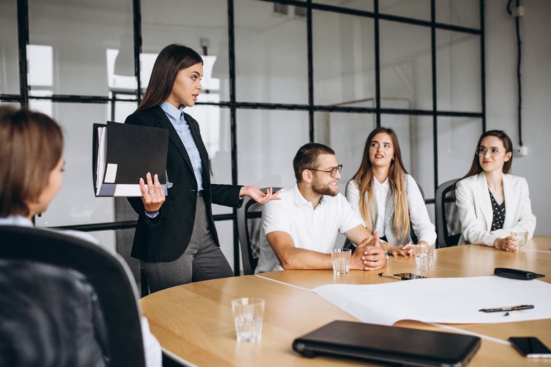Team meeting in a modern office, with a presenter standing and holding a laptop near seated colleagues.