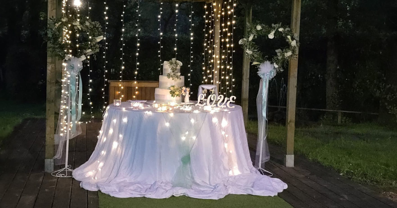 Tavolo da esterno decorato con una tovaglia bianca drappeggiata, fiori, luci e la scritta