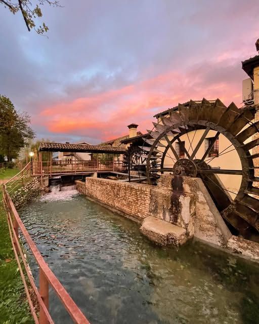 Una storica ruota idraulica accanto a un canale al tramonto, con nuvole rosa e edifici in pietra che costeggiano la riva.