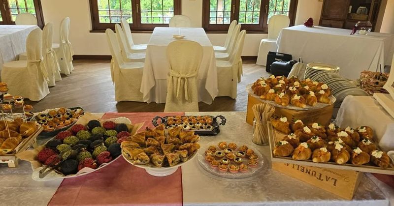 Tavolo da buffet con pasticcini, frutta e dessert in una luminosa sala da pranzo con sedie coperte.