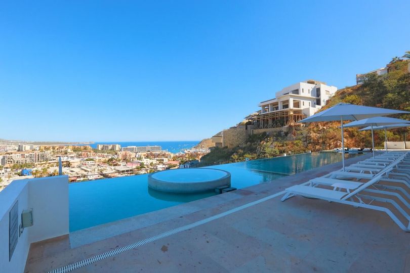 Infinity pool overlooking a coastal town and ocean under a blue sky, with white lounge chairs and a building.