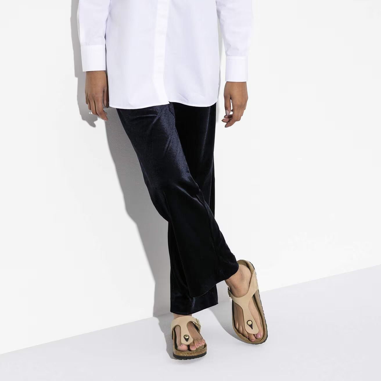 Person in a white shirt and black pants, standing with crossed legs in tan sandals against a white background