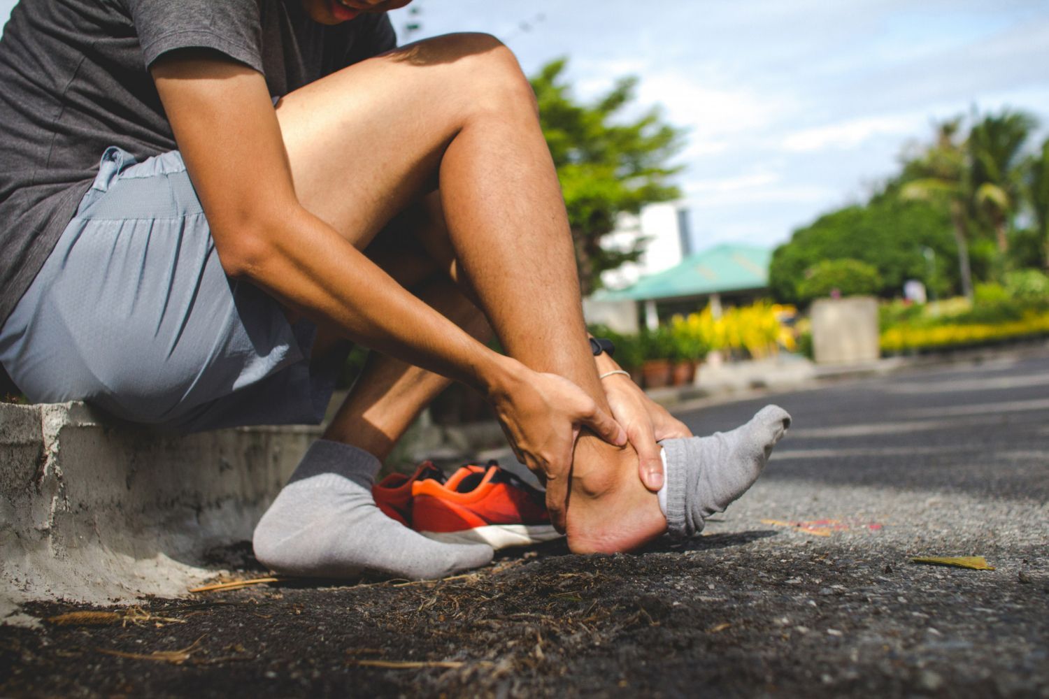 Person sitting on curb, holding ankle, appearing to be in pain. Outdoors, sunny day.