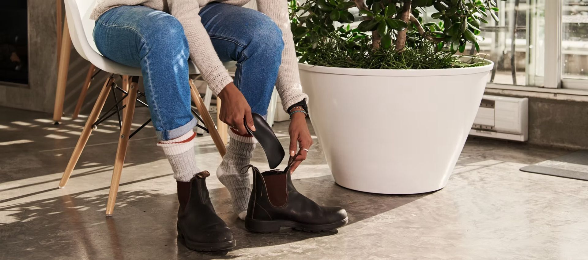 Person pulling on brown boots while seated indoors near a large white planter.
