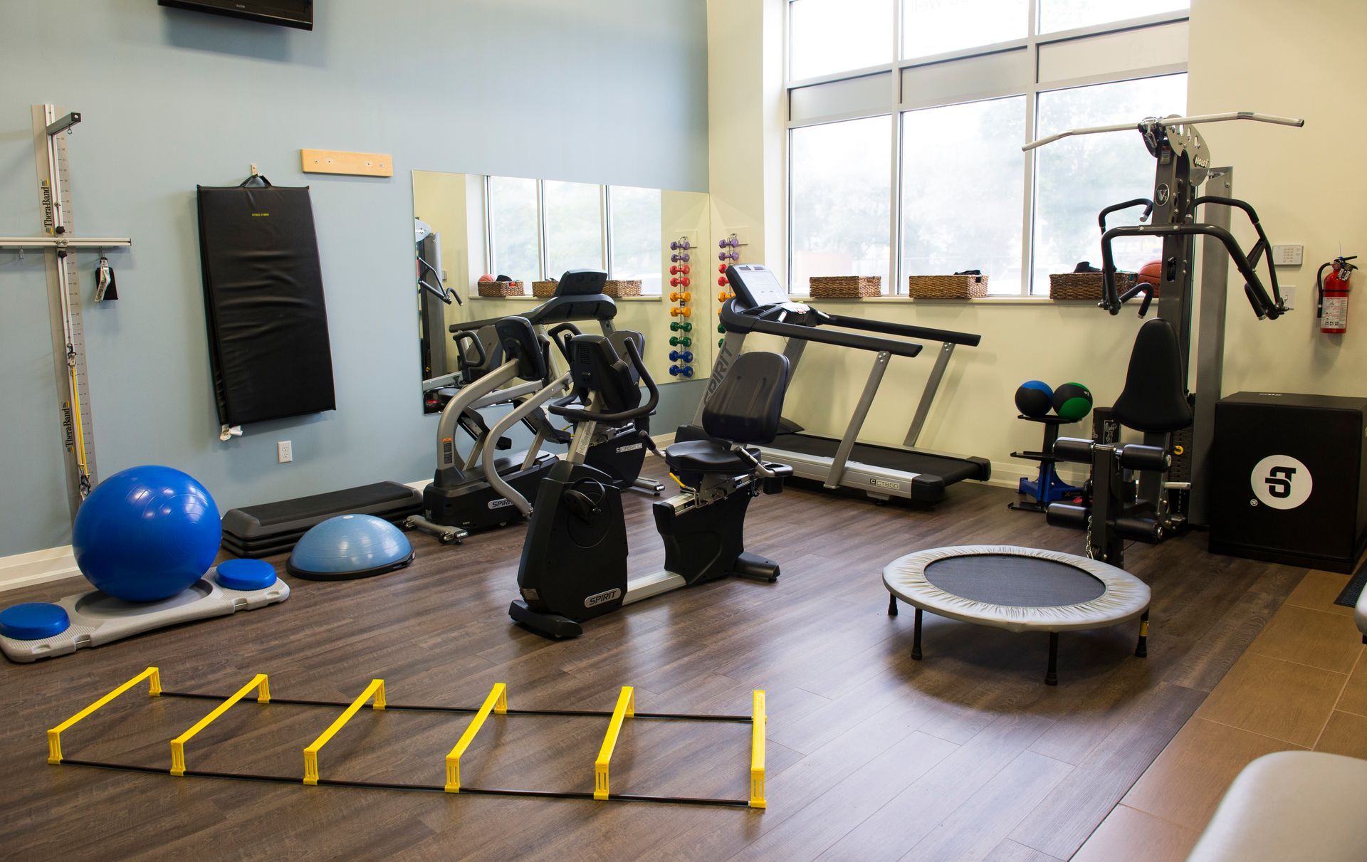 Gym room with exercise bike, treadmill, balance ball, hurdles, and trampoline on a wood floor.