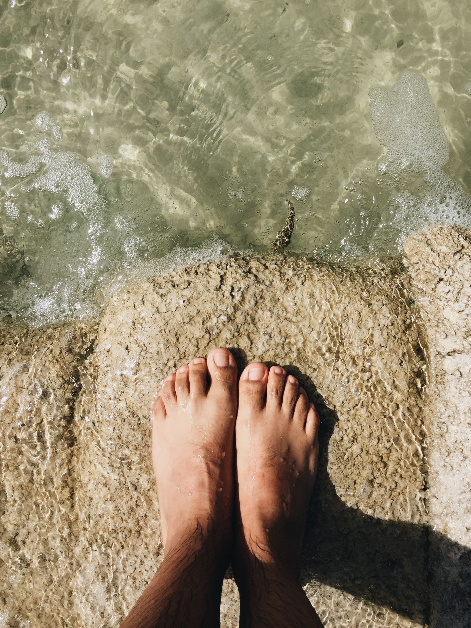 A person 's feet are standing on a rock near the water.