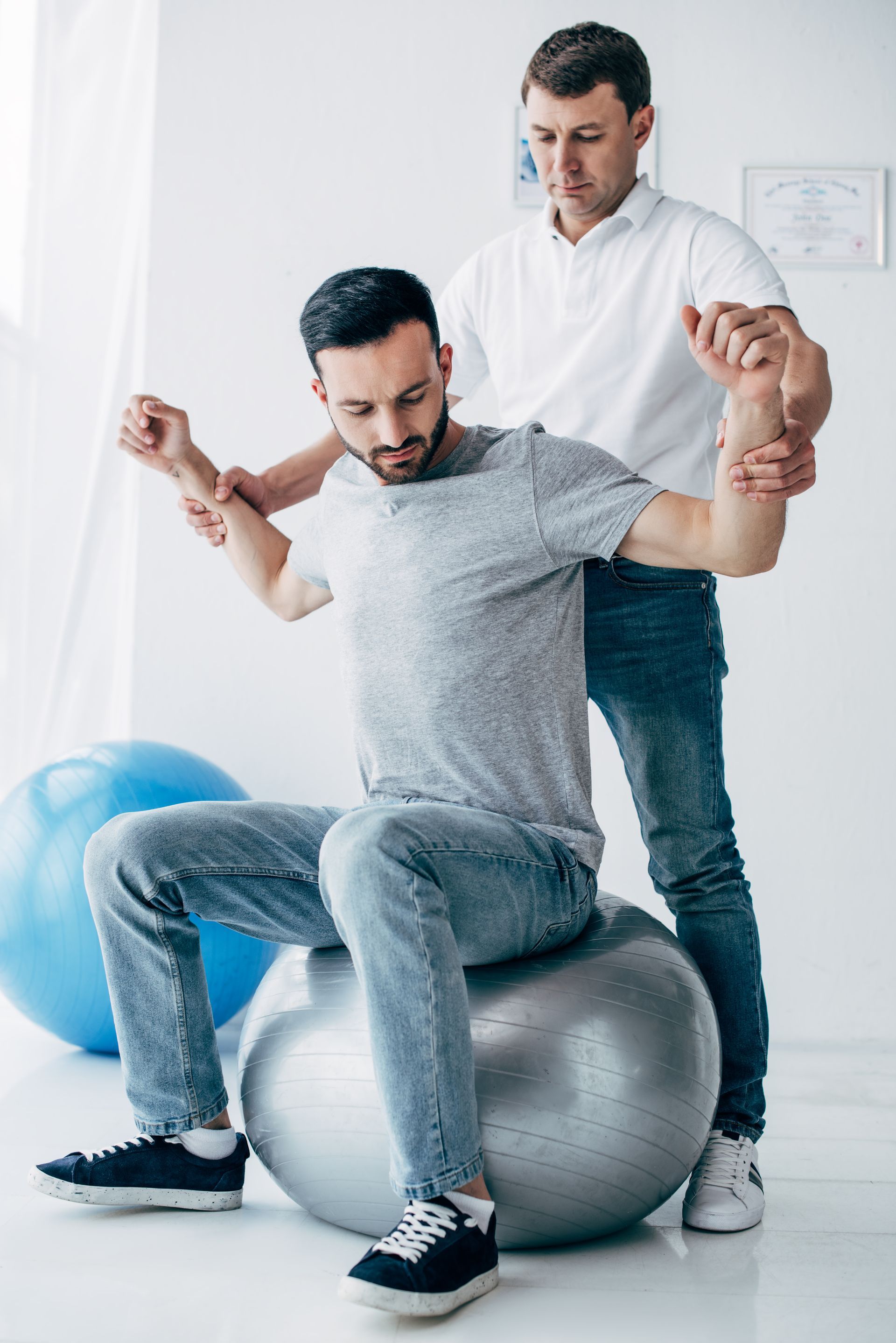 A man is sitting on an exercise ball while a man stands behind him.
